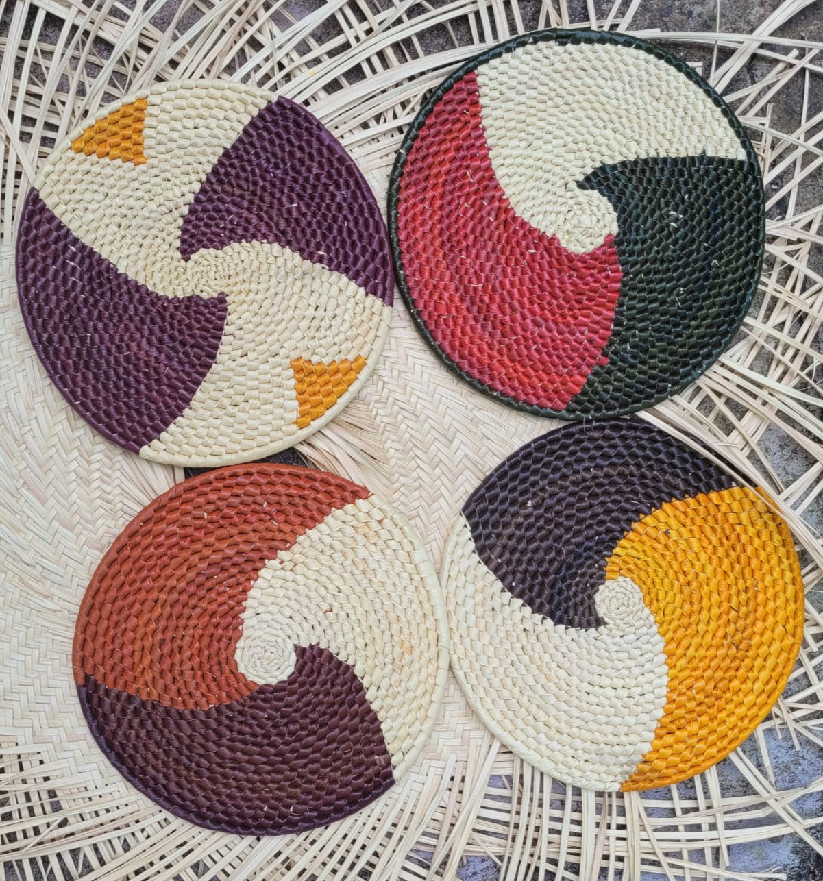 "Four circular woven trivets with swirling two-tone patterns in shades of yellow, black, white, red, green, and purple, on a herringbone wicker background.