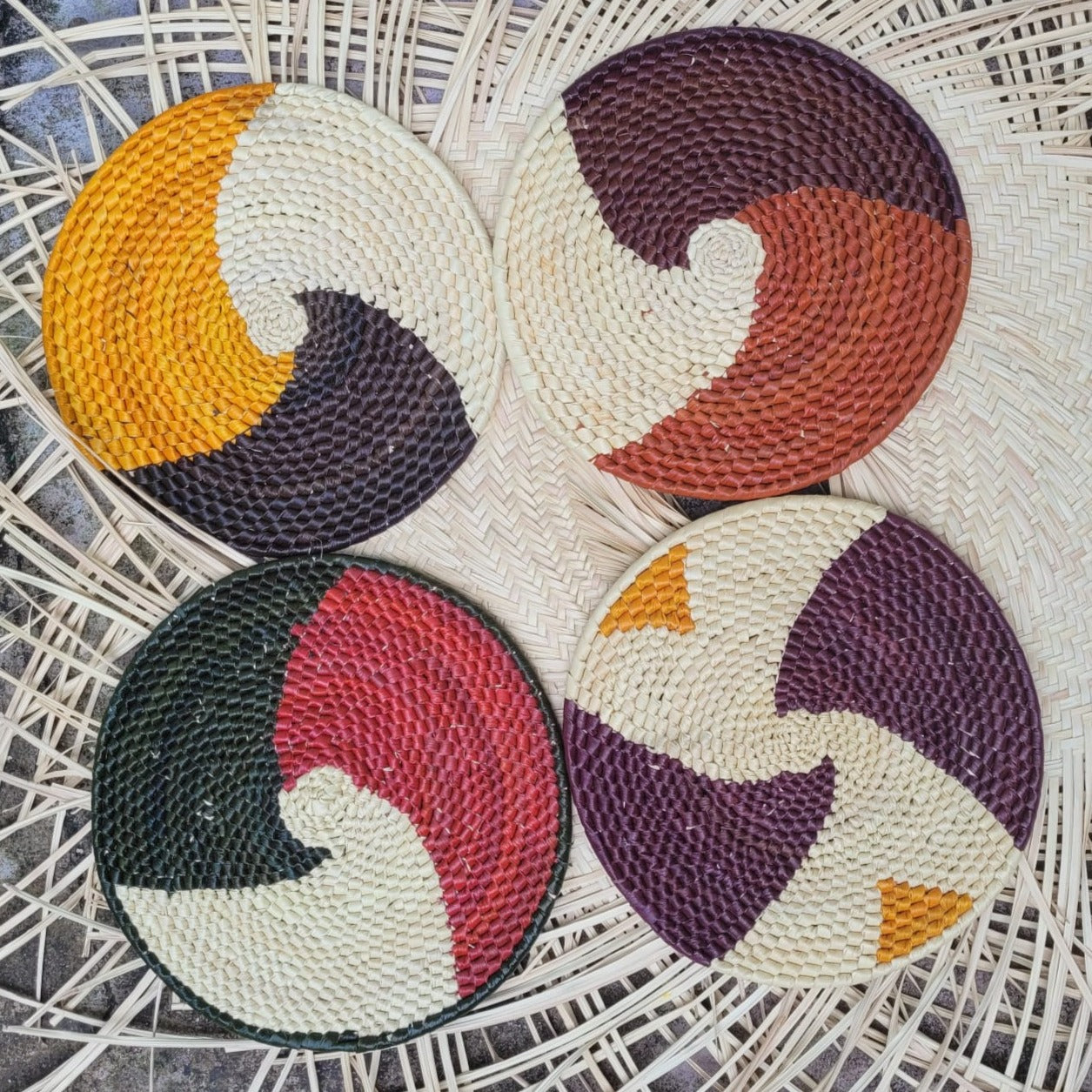 Four woven trivets with swirling two-tone patterns in yellow, white, brown, red, green, black, and purple on a herringbone wicker background.