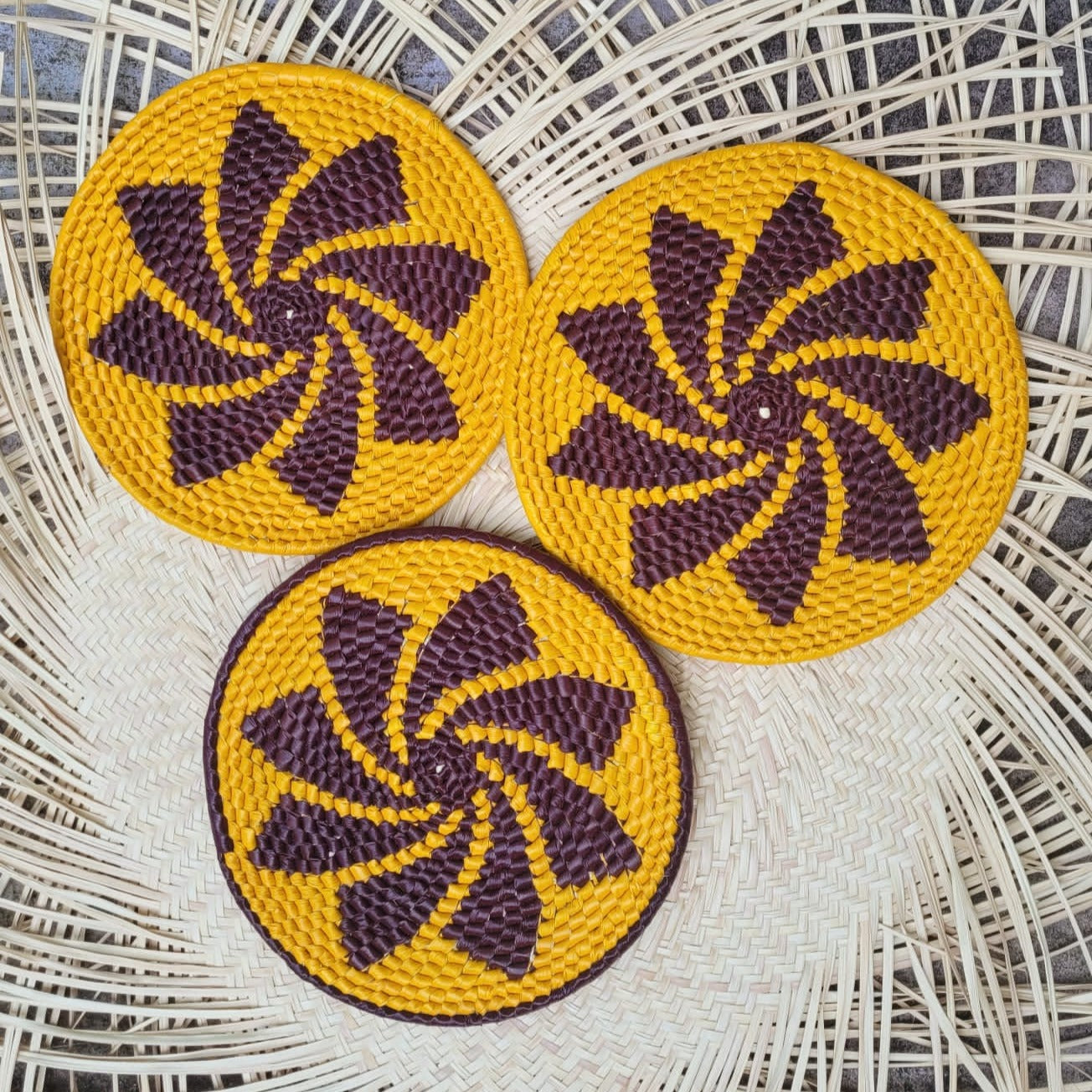 Three tucuma fiber trivets, vibrant yellow with purple spiral patterns, rest on a wicker surface. Their design evokes spinning pinwheels, set against a textured herringbone backdrop.