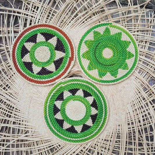 Three circular handwoven trivets crafted from tucuma fibers featuring geometric patterns in green, white, black, and brown, on a herringbone wicker surface.