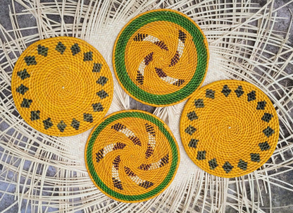 Four bright yellow woven trivets with green borders; two have a spiral pattern with brown accents, and two feature a ring of black diamonds on a wicker background.