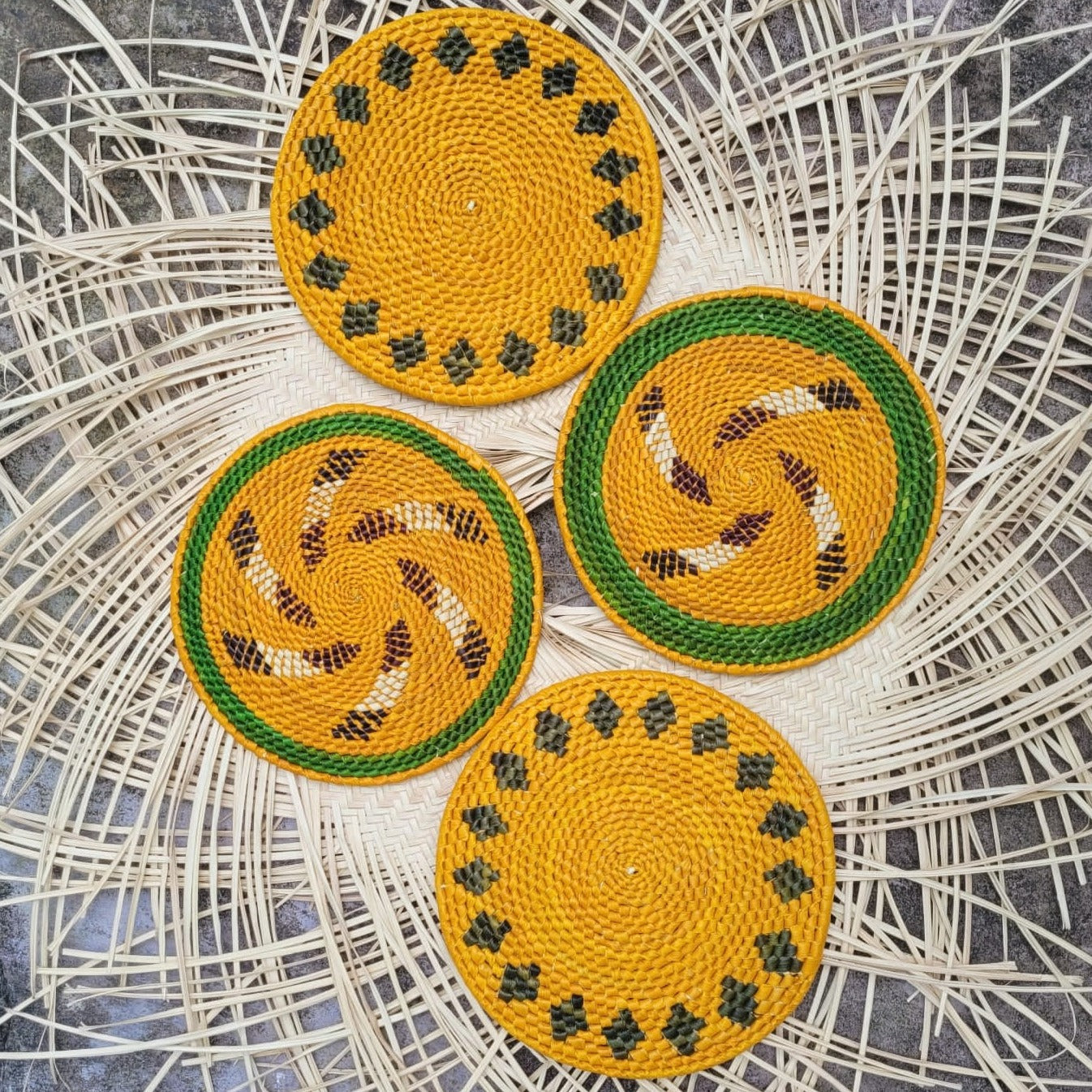 Four yellow handwoven trivets with green edges on a wicker background, two with brown and cream spiral accents, two with black dot patterns.