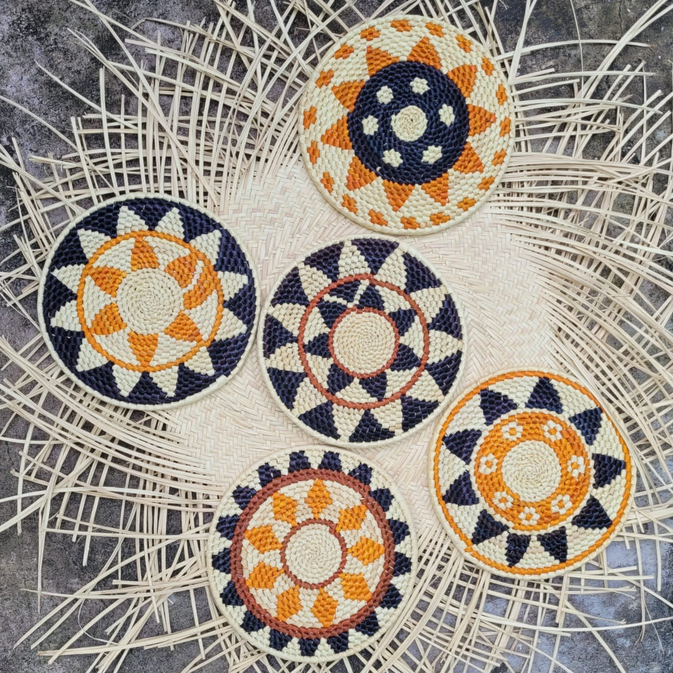 Four handcrafted trivets with intricate geometric patterns in navy blue, white, and orange hues, laid out on a natural herringbone-patterned wicker background.