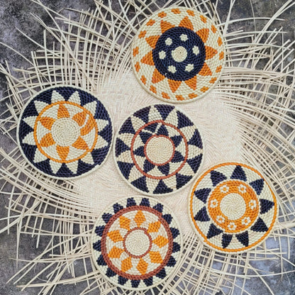 Four handcrafted trivets with intricate geometric patterns in navy blue, white, and orange hues, laid out on a natural herringbone-patterned wicker background.