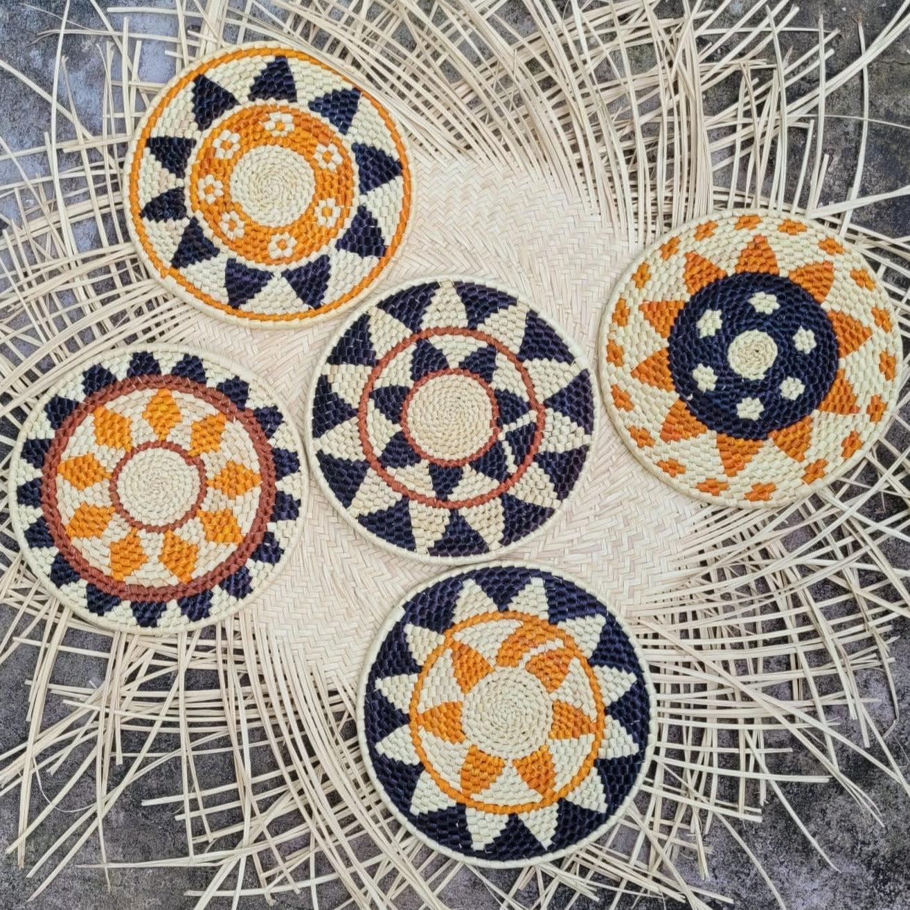 Five handcrafted trivets with orange, navy, and white geometric patterns laid out on a textured herringbone wicker mat.