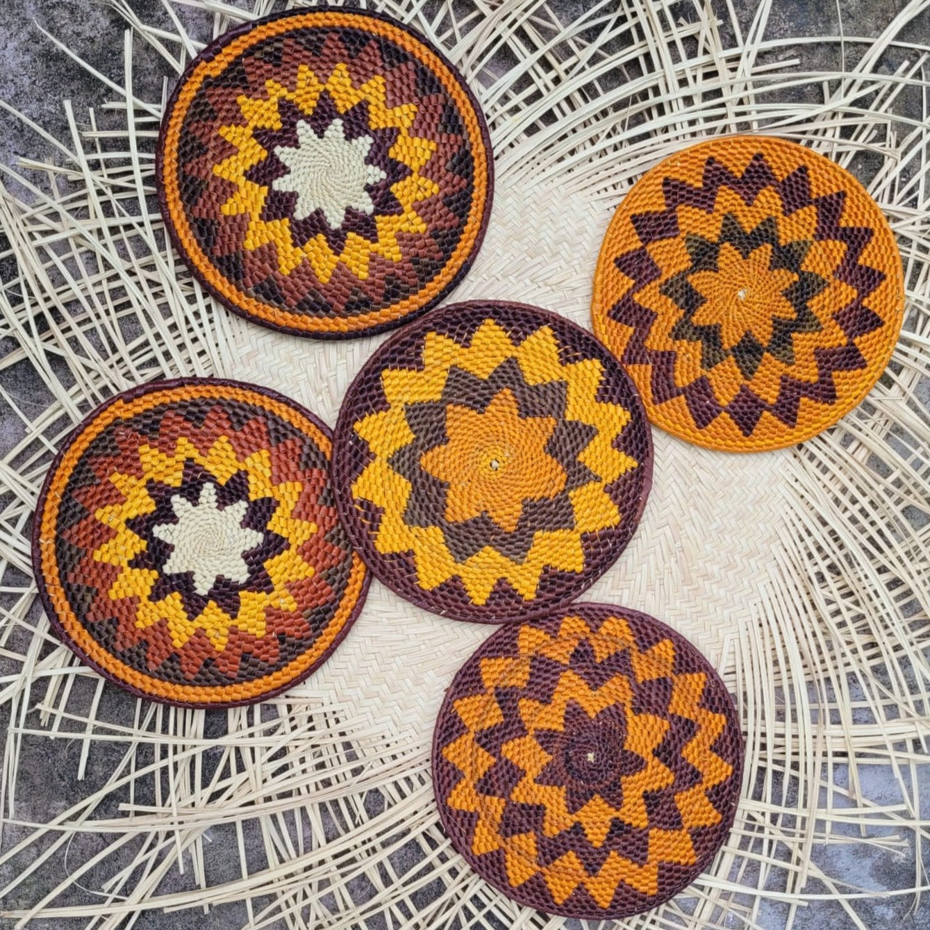  Five round woven trivets on a herringbone mat with starburst patterns in orange, yellow, and dark brown. The intricate designs and colors evoke a warm, natural aesthetic.