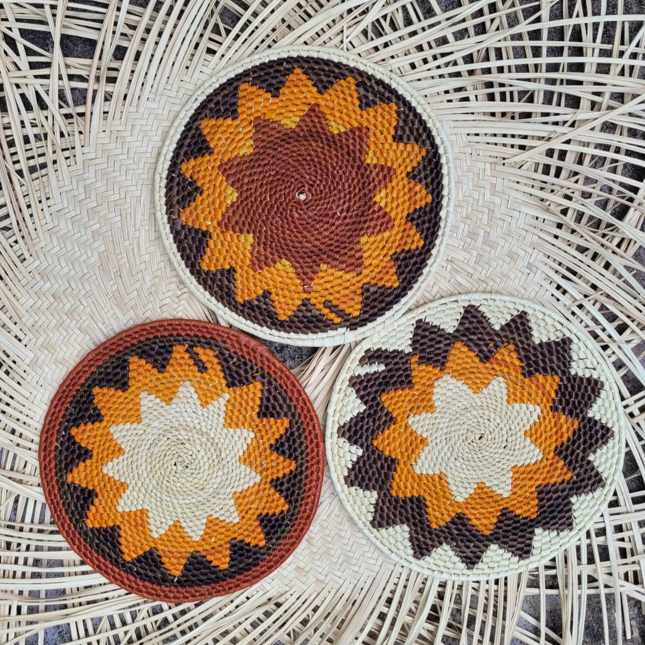 Image of three handwoven tucuma trivets with starburst patterns in orange, brown, and white hues on a herringbone wicker backdrop.