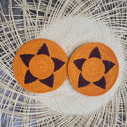 The image displays two handwoven trivets with a central orange spiral design, embellished with a dark purple, four-pointed star pattern extending towards the edge.