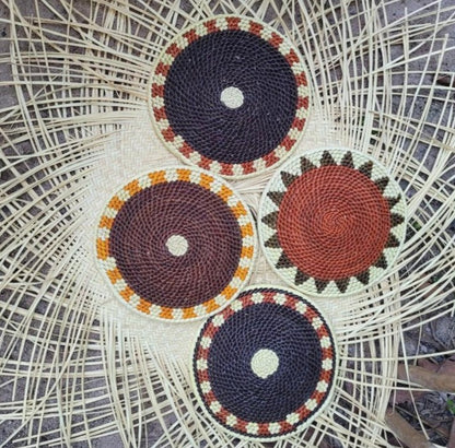 Four handwoven trivets on a wicker surface: two in navy with a central circle, one in shades of brown with a spiral pattern, and one in navy with an orange and cream checkered border.