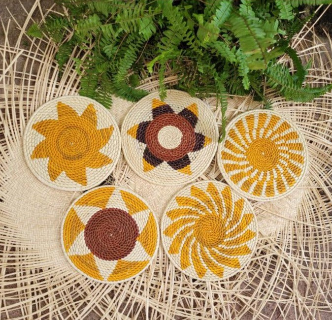 Five handwoven trivets featuring radiant patterns in yellow, cream, and brown, set against a herringbone wicker backdrop with green fern leaves.