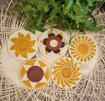 Five handwoven trivets featuring radiant patterns in yellow, cream, and brown, set against a herringbone wicker backdrop with green fern leaves.