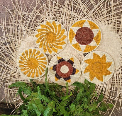  Five handwoven trivets with dynamic patterns in shades of yellow, brown, and cream are arranged on a wicker background alongside verdant fern leaves.