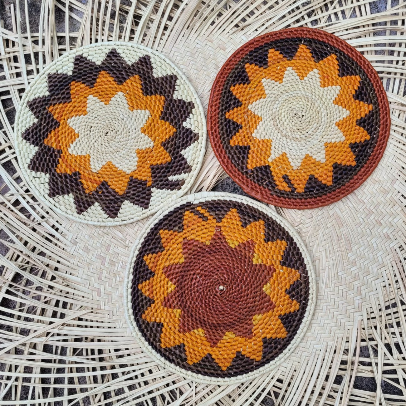 Three handwoven tucuma trivets on a wicker background, with sunburst patterns in brown, orange, and cream.