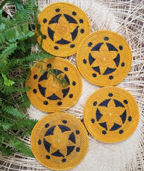 Five yellow handwoven trivets, each with a unique black geometric pattern, displayed on a textured wicker background with green fern leaves adding a touch of nature.