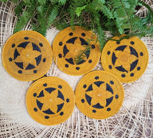 Five vibrant yellow handwoven trivets, each with a circular swirl of black accents, set against a wicker backdrop with green fern leaves.