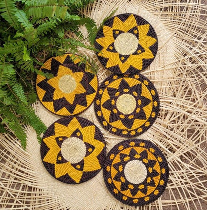 A set of five black and yellow handwoven trivets with assorted patterns, including stars and spirals, resting on a wicker mat beside fern leaves.