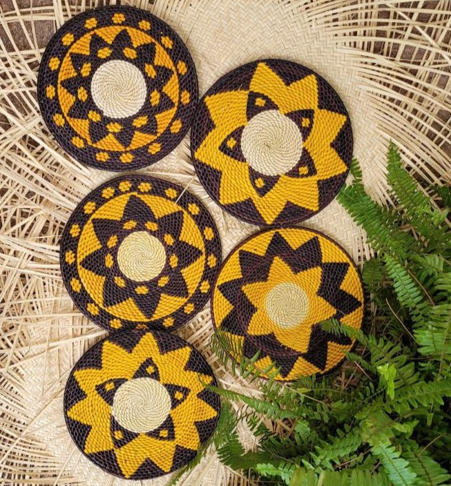 A collection of five handwoven trivets in yellow and black patterns, nestled amongst fern leaves on a woven rattan background. Each trivet features a distinct, intricate design, radiating a traditional craft aesthetic.