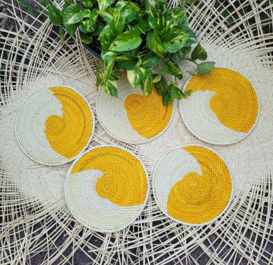 Five handwoven trivets in a cream and yellow swirl pattern, resembling stylized suns, set against a herringbone wicker backdrop with a lush green plant to the side.