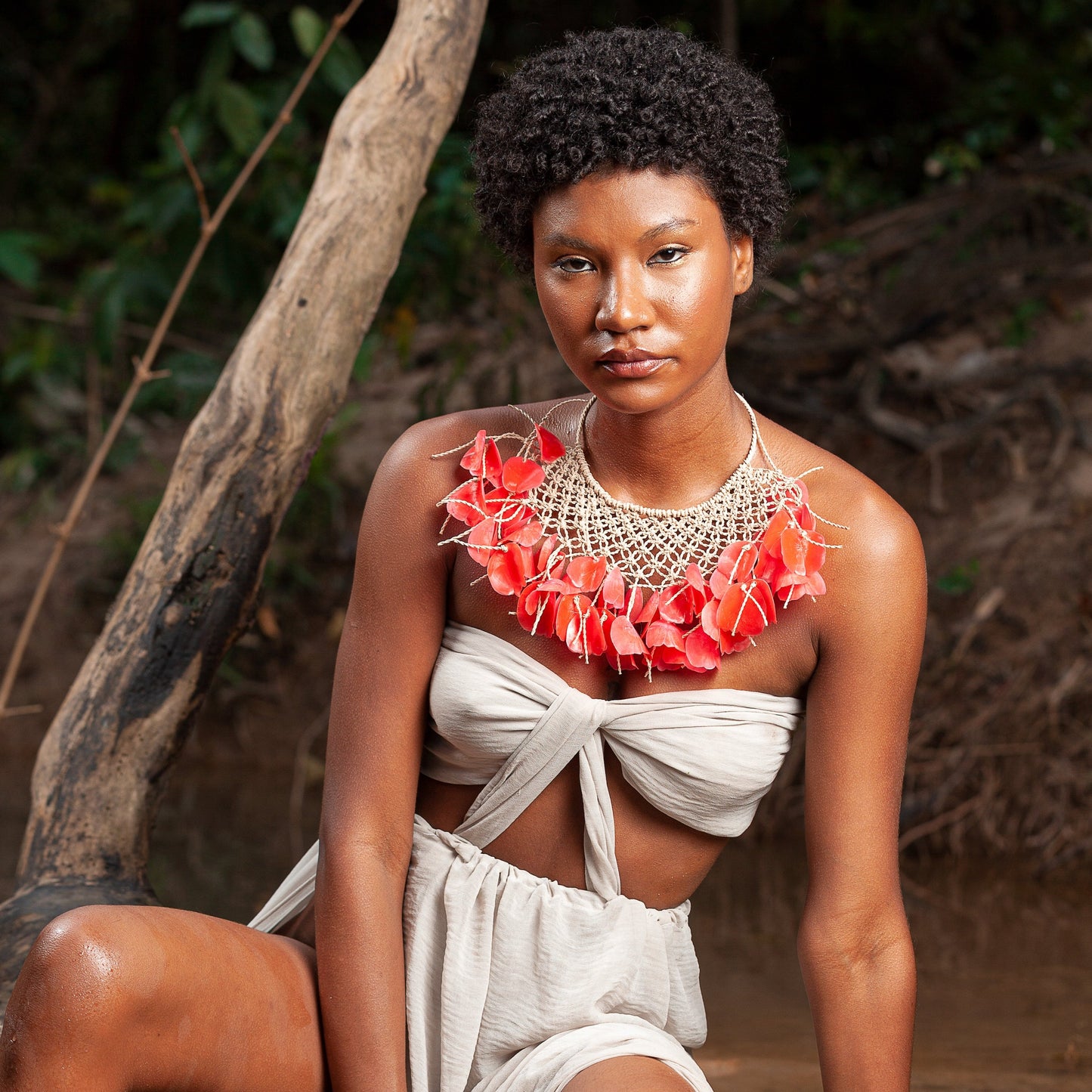 The image shows a model seated outdoors, wearing a striking necklace crafted with miriti fiber and vibrant red tarpon fish scales, paired with a casual, knotted top. Her confident gaze and natural beauty are emphasized by the unique, handcrafted jewelry.