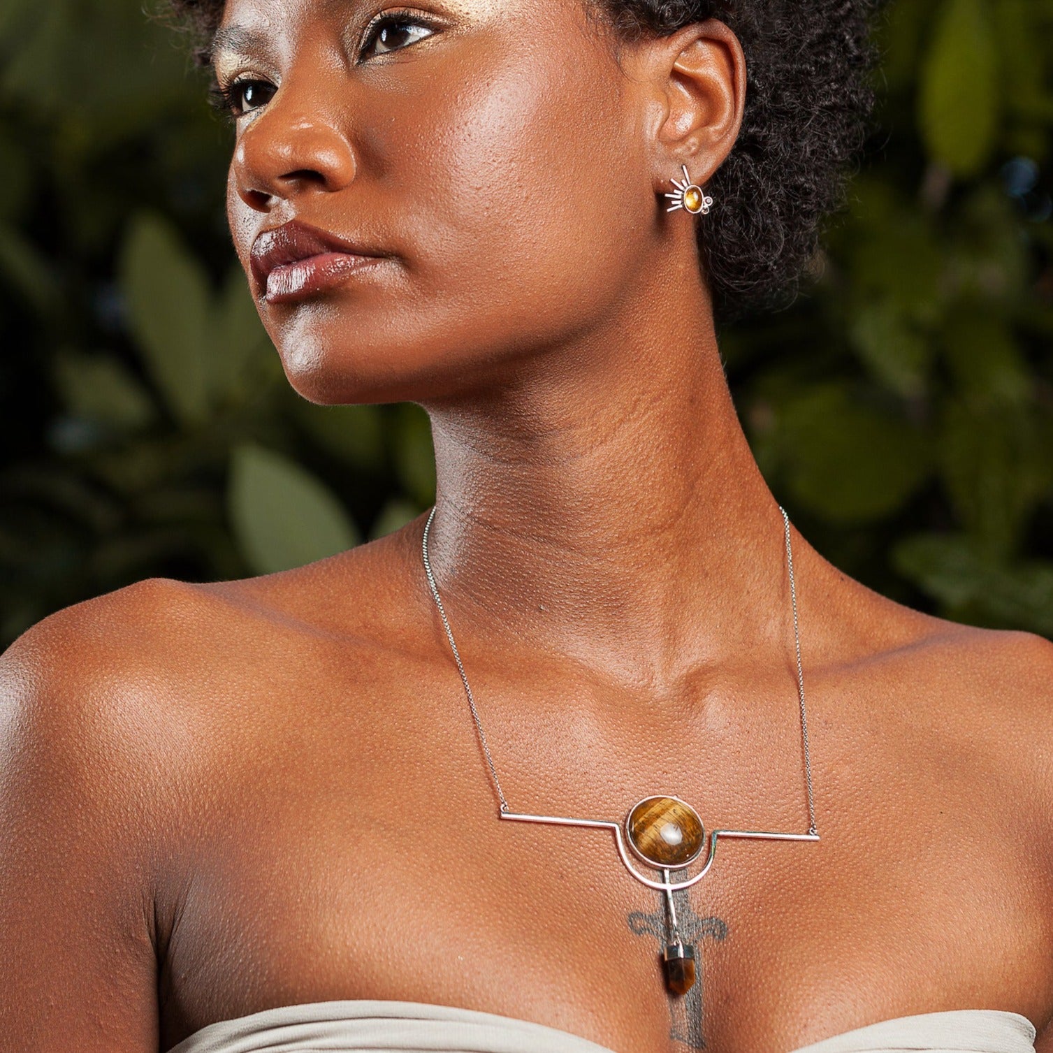 An elegant woman models a sterling silver necklace, featuring a prominent tiger's eye gemstone centerpiece, with complementary earrings, set against a leafy backdrop