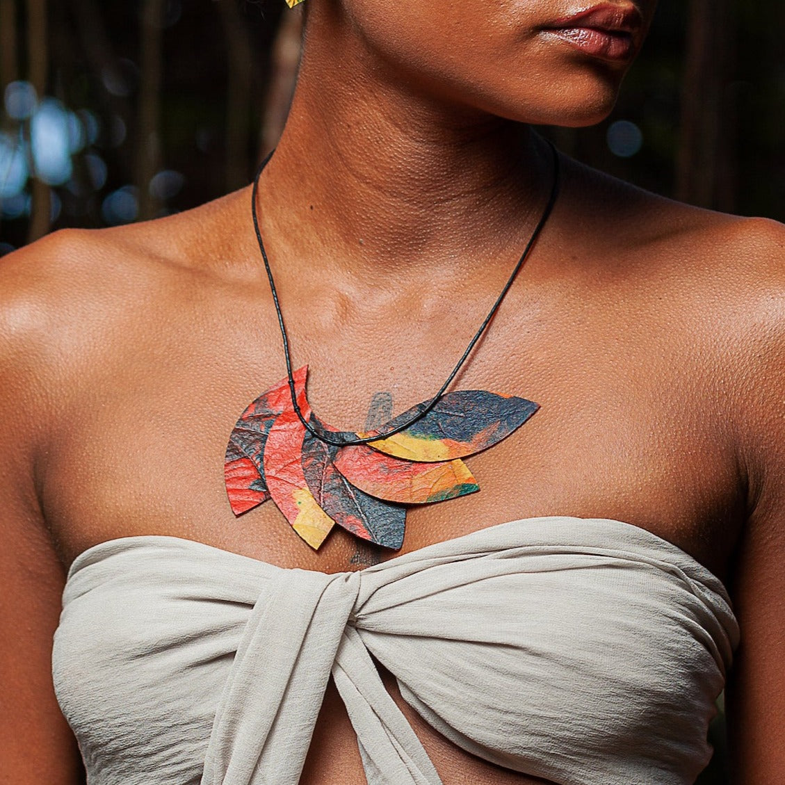 Model wears handmade necklace with leaf-shaped pendants crafted from natural rubber & açaí seed fiber. Autumnal colors & intricate vein patterns evoke organic beauty.