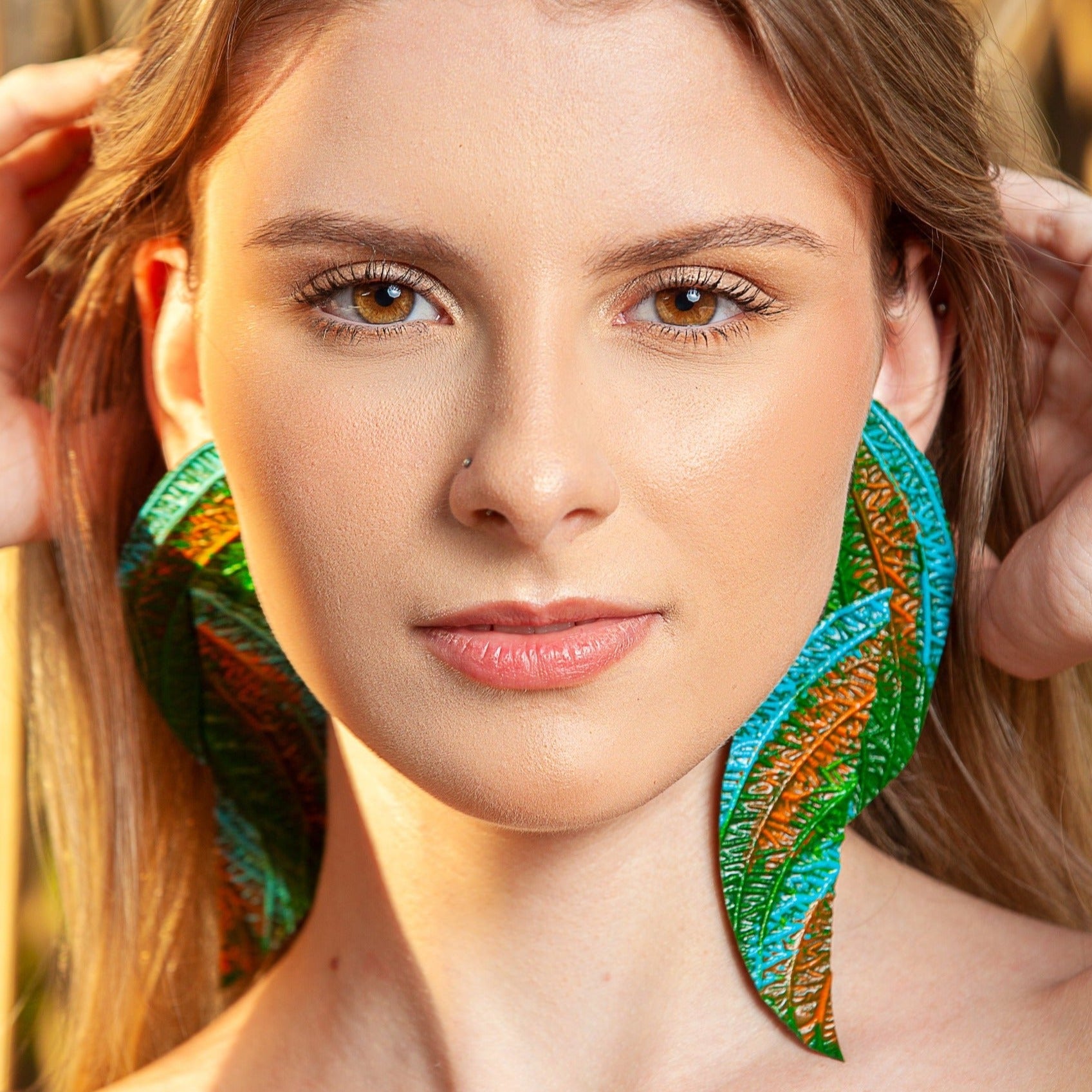 A model wearing large leaf-shaped earrings, made from rubber latex and açaí fibers by artisans, with intricate green, blue and orange patterns.