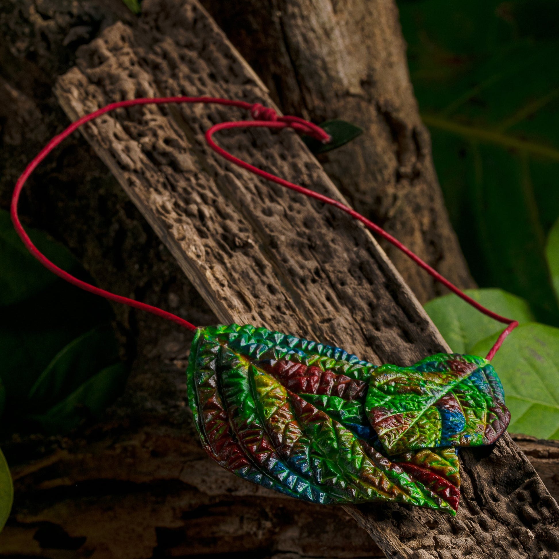 Colorful rubber leaf necklace on a red açaí fiber cord, set against a tree bark backdrop, showcasing rich textures and vibrant hues.