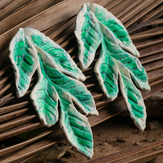 Handmade leaf-shaped earrings on a woven surface, crafted from sustainable rubber latex and açaí fibers by local artisans, in varied green shades.