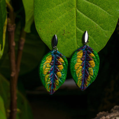 Natural rubber leaf-shaped earrings with vibrant green, yellow, and purple hues, accented with açaí seed fiber, hang before a backdrop of real leaves, showcasing the Amazon's essence, nickel-free.