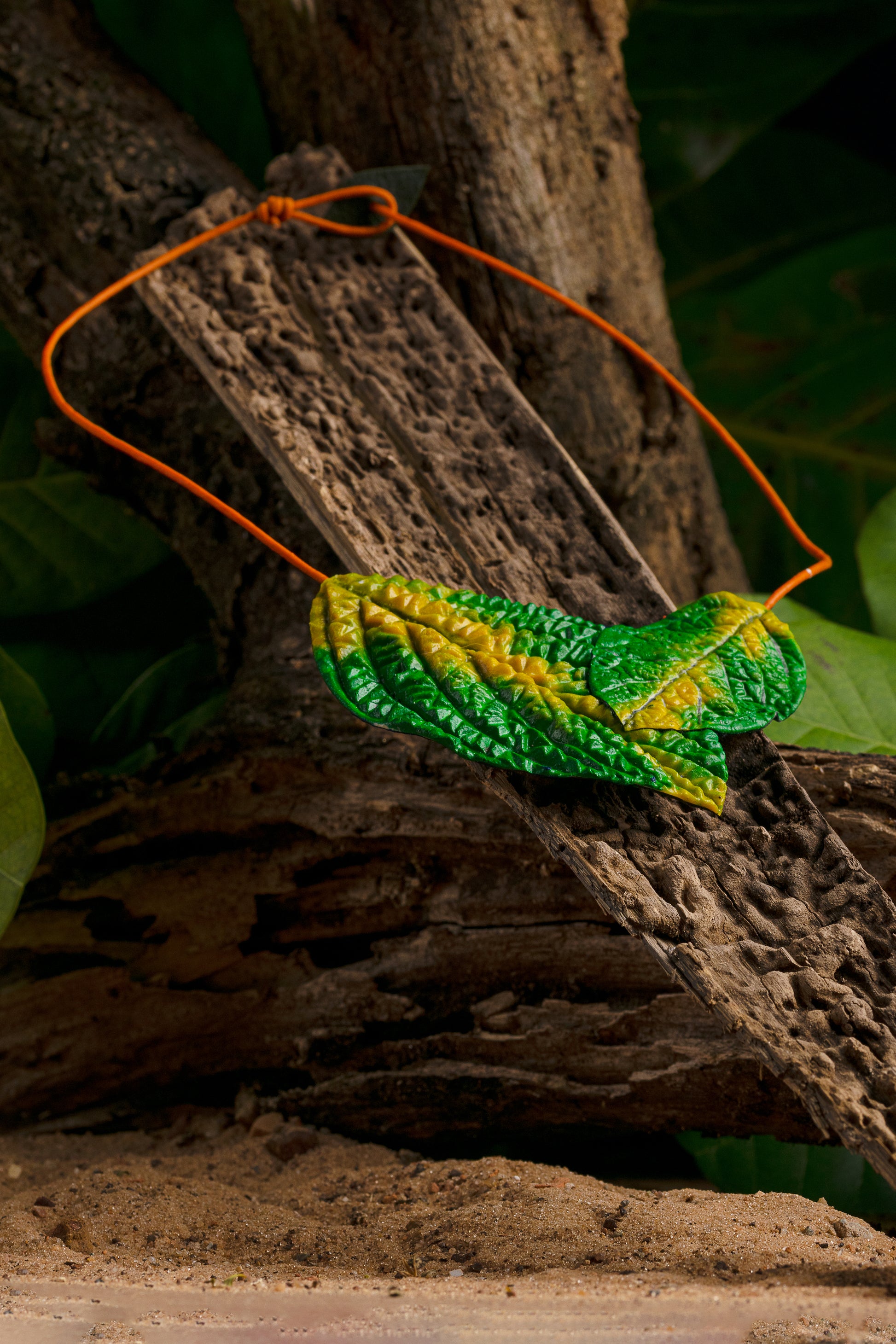 Artisan necklace with vibrant green rubber leaves on an orange açaí fiber cord, draped over a textured tree trunk amidst lush foliage