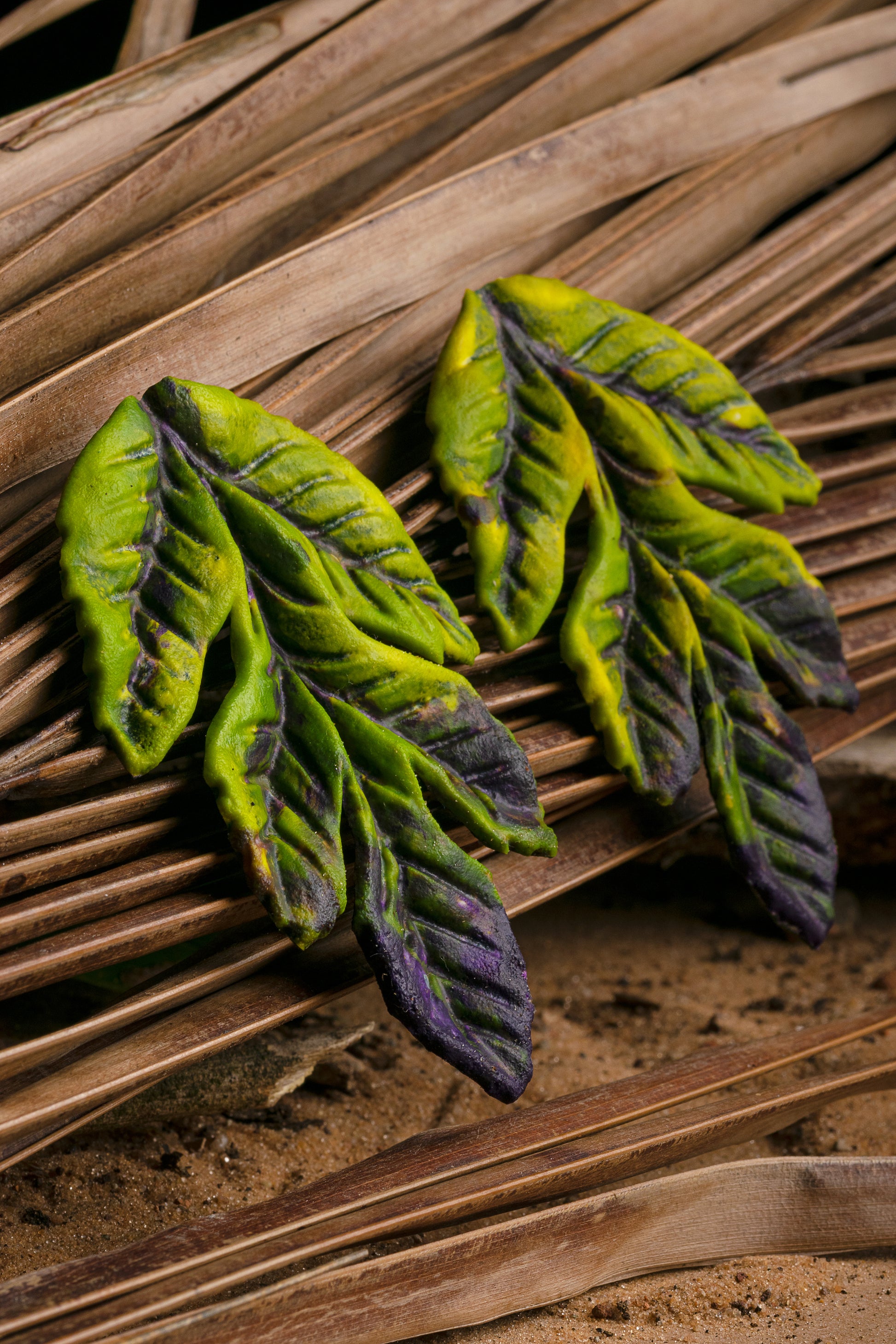 Leaf earrings with green to purple gradient, crafted from rubber latex and açaí fibers by artisans, on a natural mat.