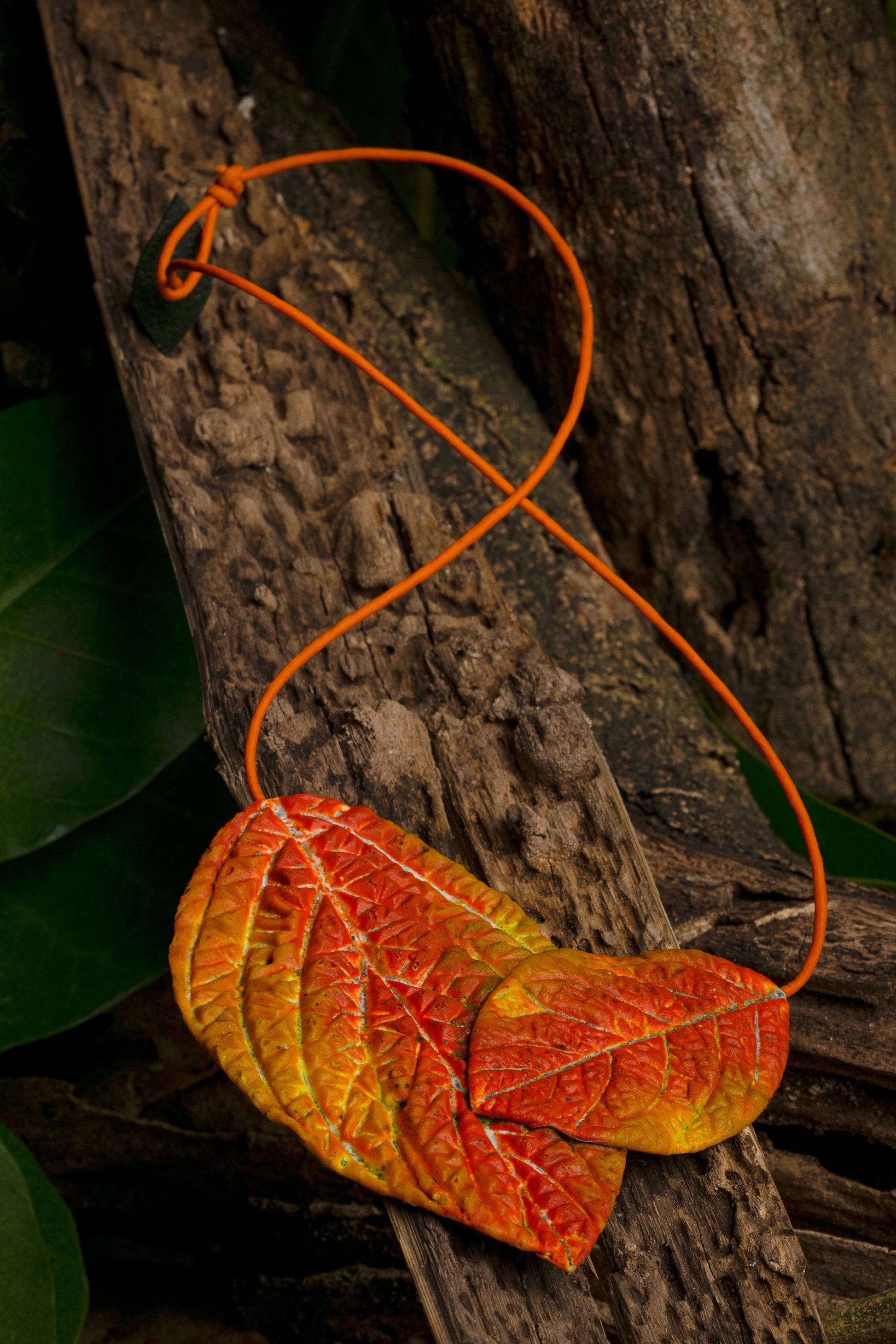 A necklace of rubber leaves in maroon, orange, and yellow hues on an orange açaí fiber cord, laid on wood amidst green leaves, showcasing artisanal craft