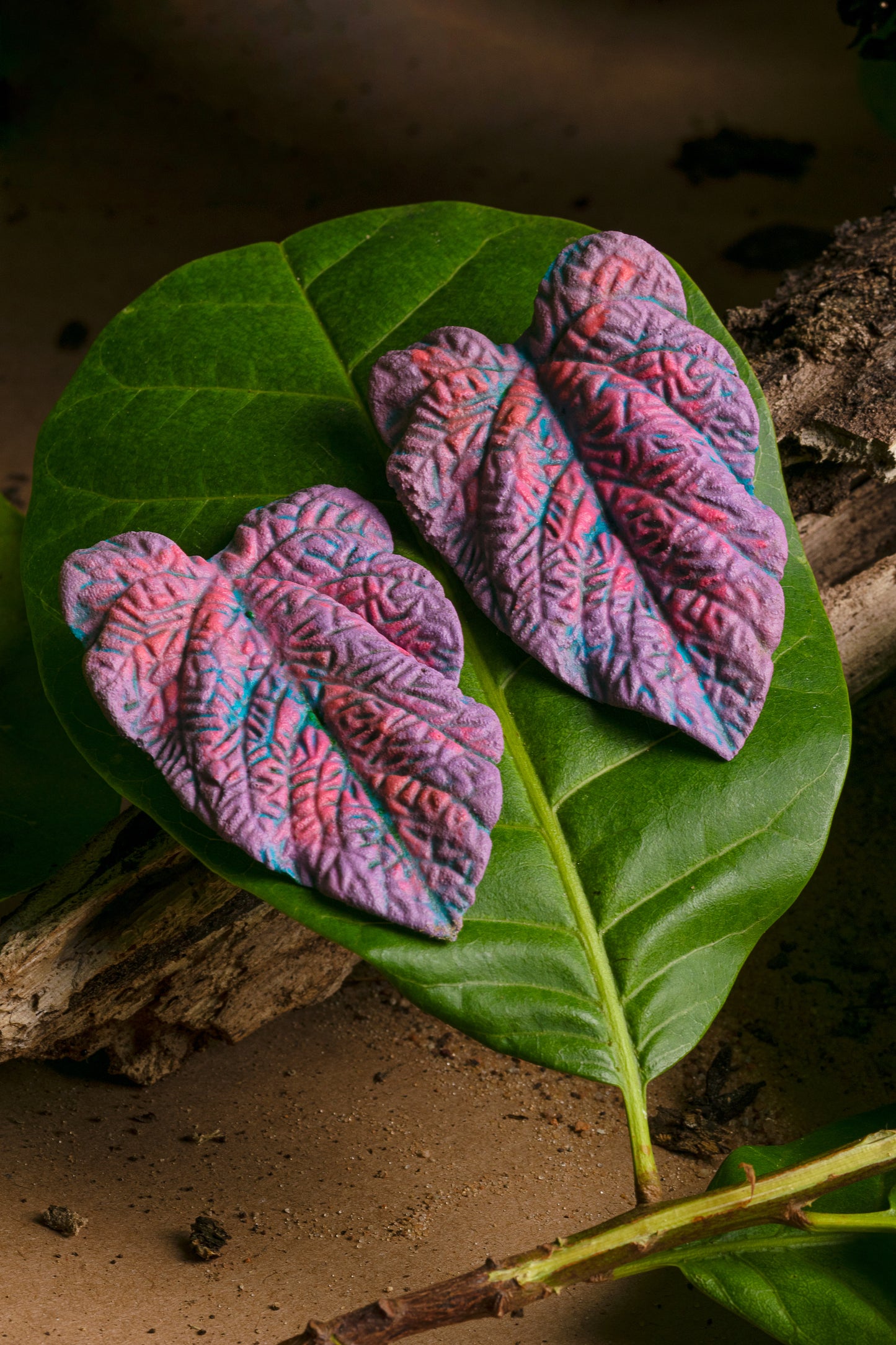 Handcrafted nickel-free earrings. Vibrant blue and purple leaf design with intricate veins, made from natural rubber and açaí seed fiber. Resting on a green leaf. 