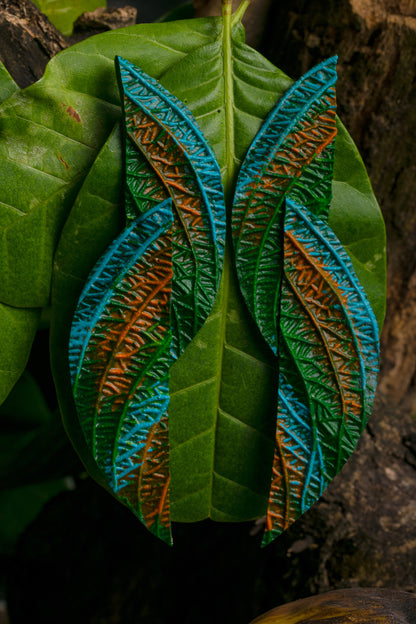 Vibrant green, blue, and orange leaf earrings dangle from a lush rainforest leaf backdrop.