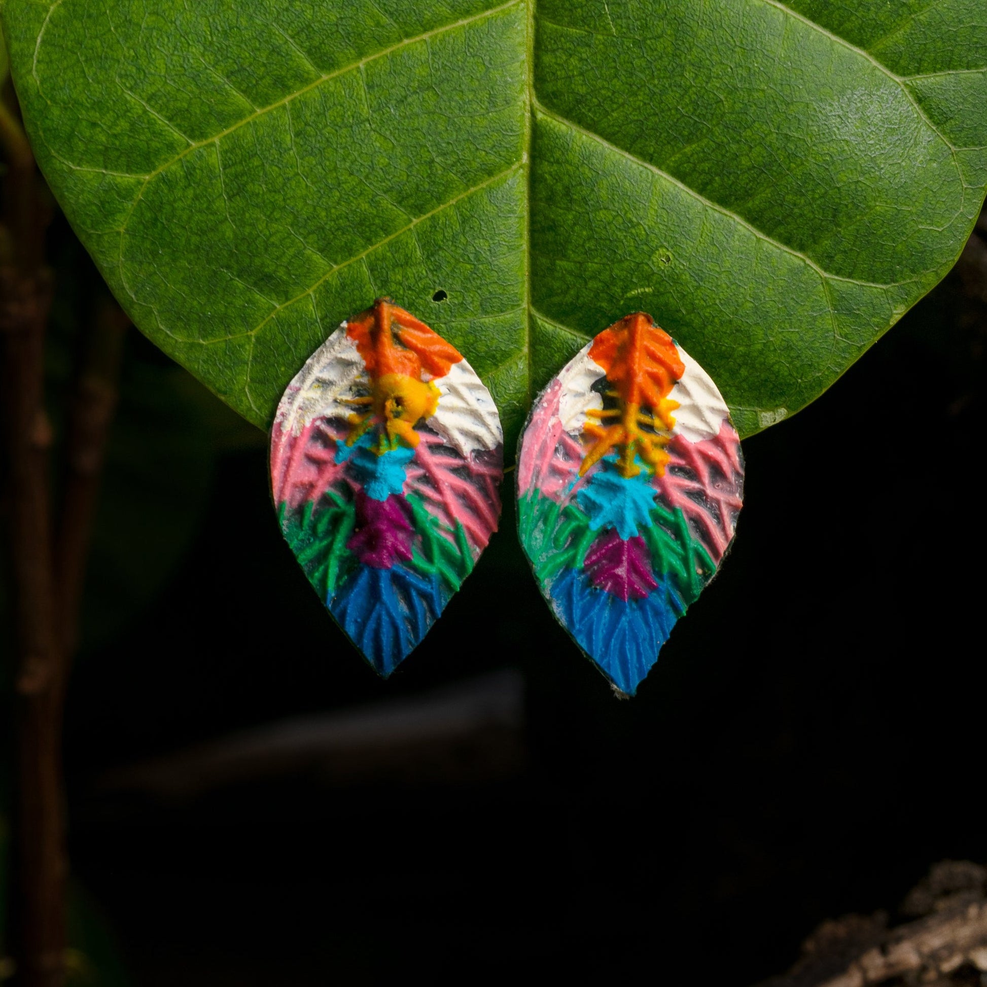 Colorful leaf-shaped earrings with a rainbow of hues, from orange to blue, on natural backgrounds, dangle delicately against a green leaf, reflecting the vibrancy of the Amazon.