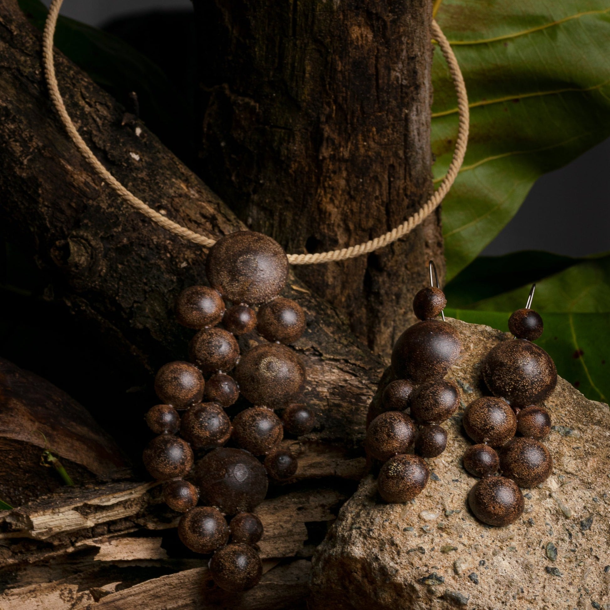 Handmade brown jewelry set, with necklace and earrings made of açaí, resin, and fiber, displayed on natural wood and stone.