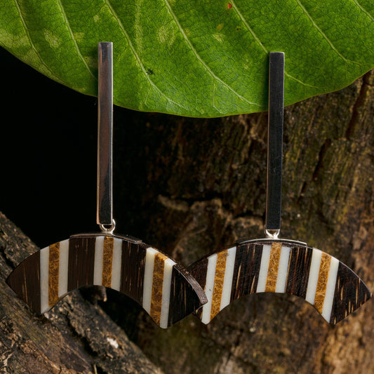 Silver comma-shaped earrings with marquetry bone inlay, hanging from a leaf against wood.