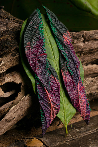 Handmade leaf earrings with vibrant purple, green, and salmon colors on a wooden backdrop. Crafted from rubber latex and açaí by artisans.