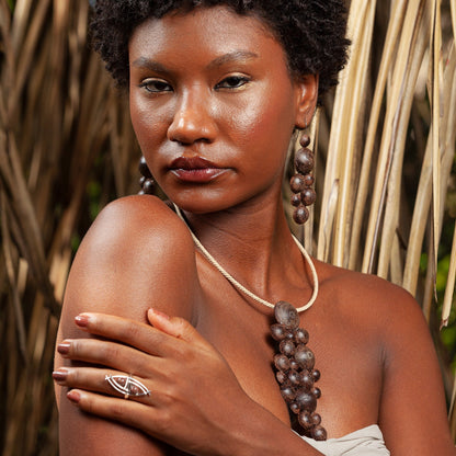 A woman with golden makeup, wearing a handmade açaí, resin, and fiber necklace and earrings set, poses while looking at the camera, showing a silver ring handmade with açaí.