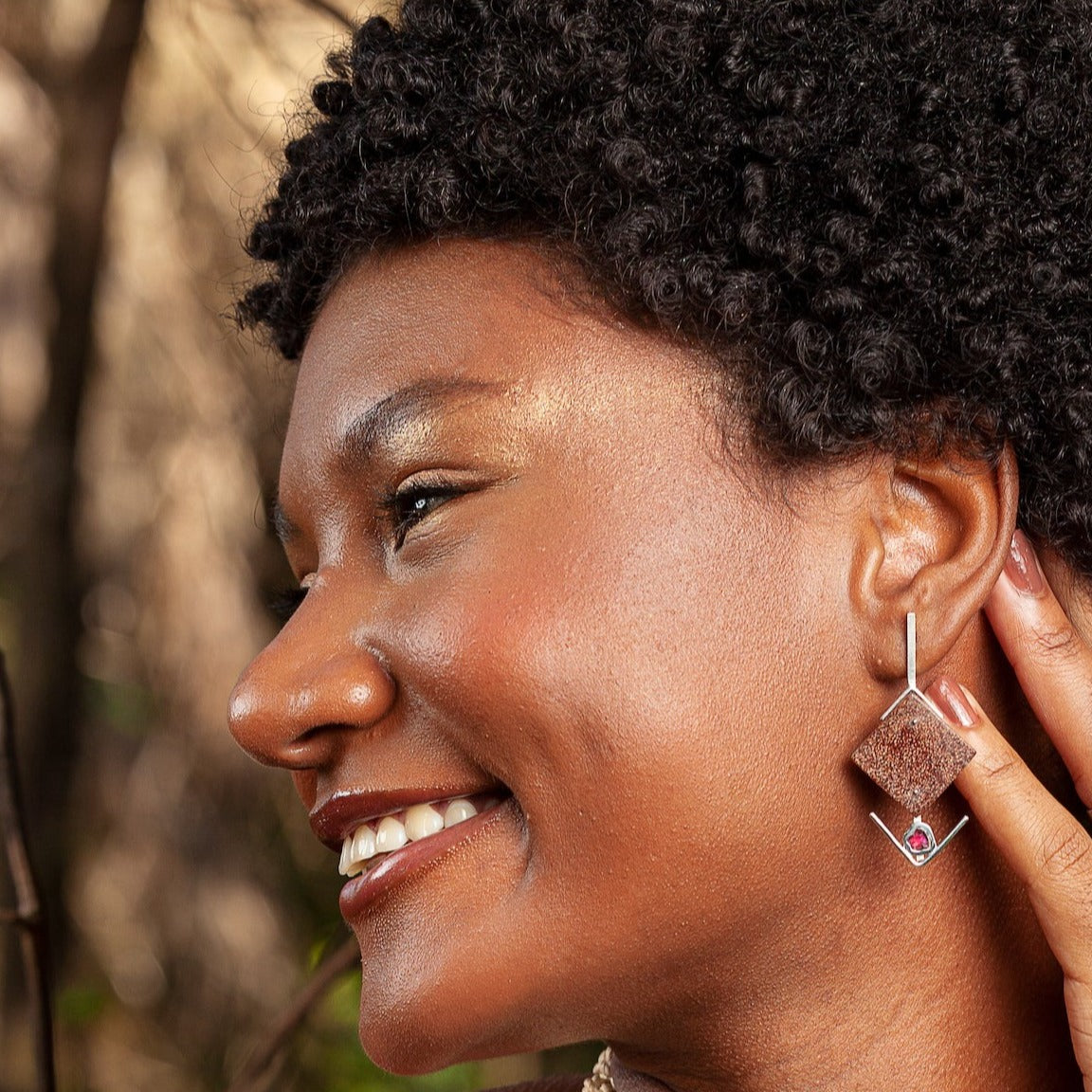 A joyful woman wears earrings made from 925 sterling silver with an amethyst gemstone, set against a base of recycled açaí and peach palm.