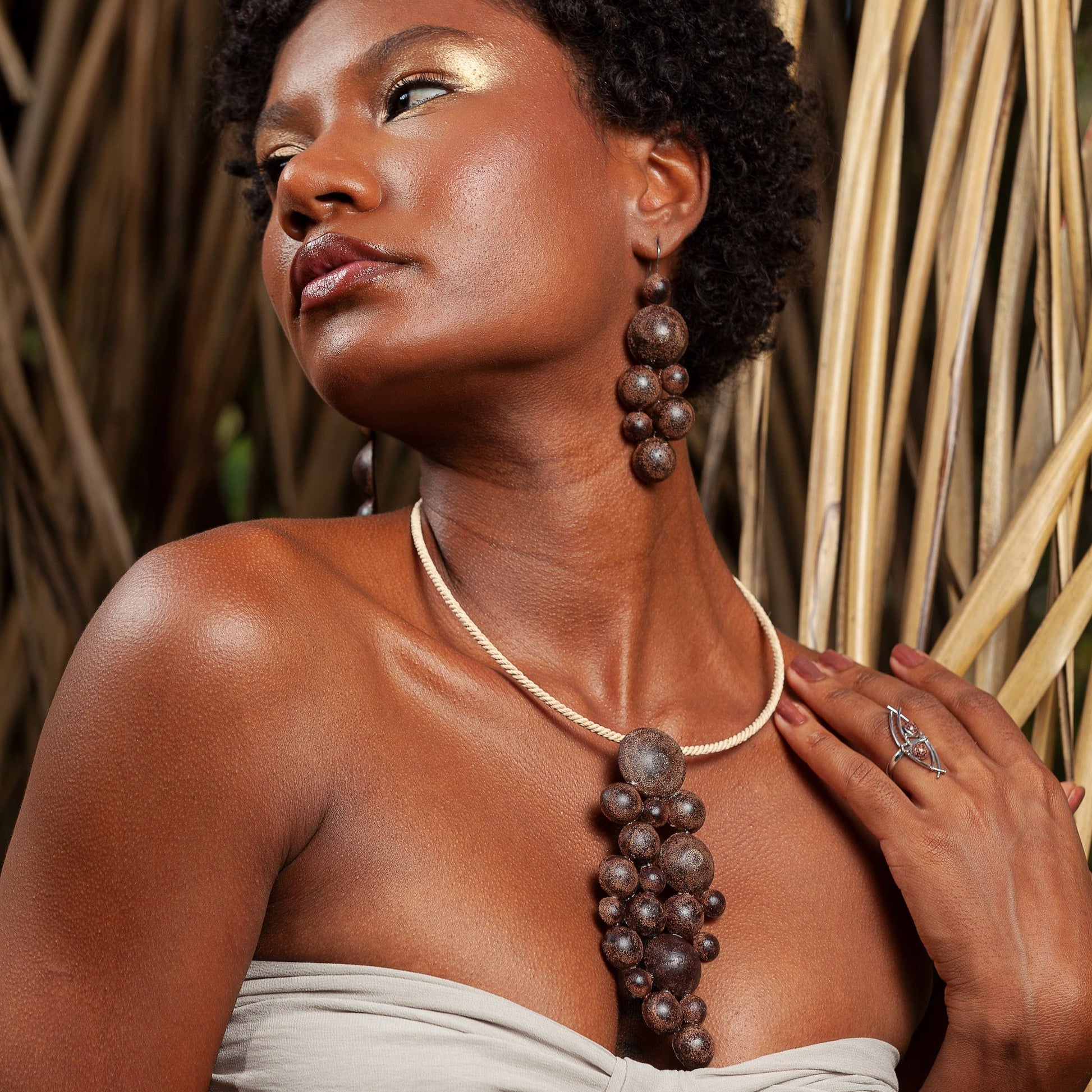 Woman with curly hair and golden makeup, wearing a handmade jewelry set crafted from açaí, resin, and fiber, posing among reeds.