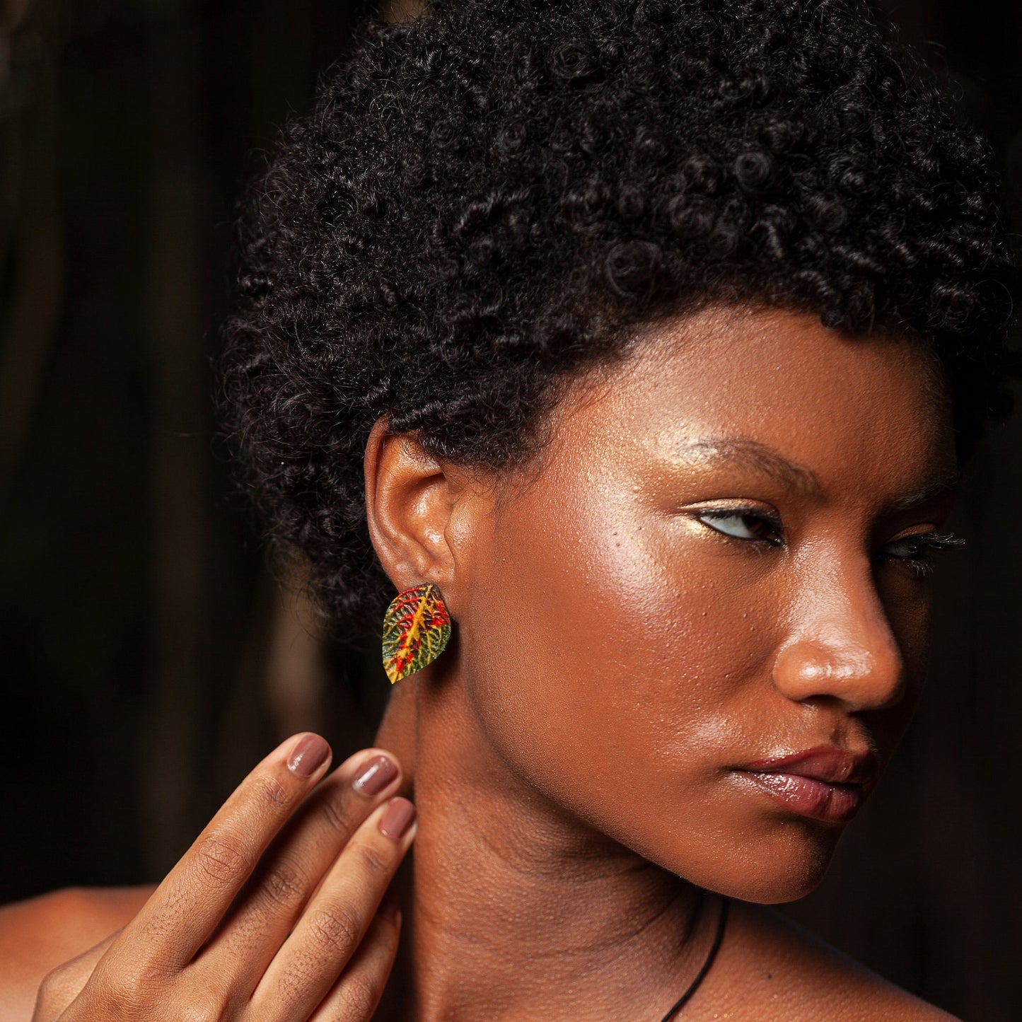 A woman with a striking profile, shimmering golden makeup, and curly hair wears an intricate leaf-shaped earring with a vivid red and green pattern, embodying the Amazon's spirit.