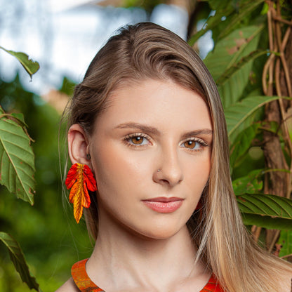 Woman with natural makeup wearing vibrant orange leaf-shaped earrings, outdoors surrounded by greenery.