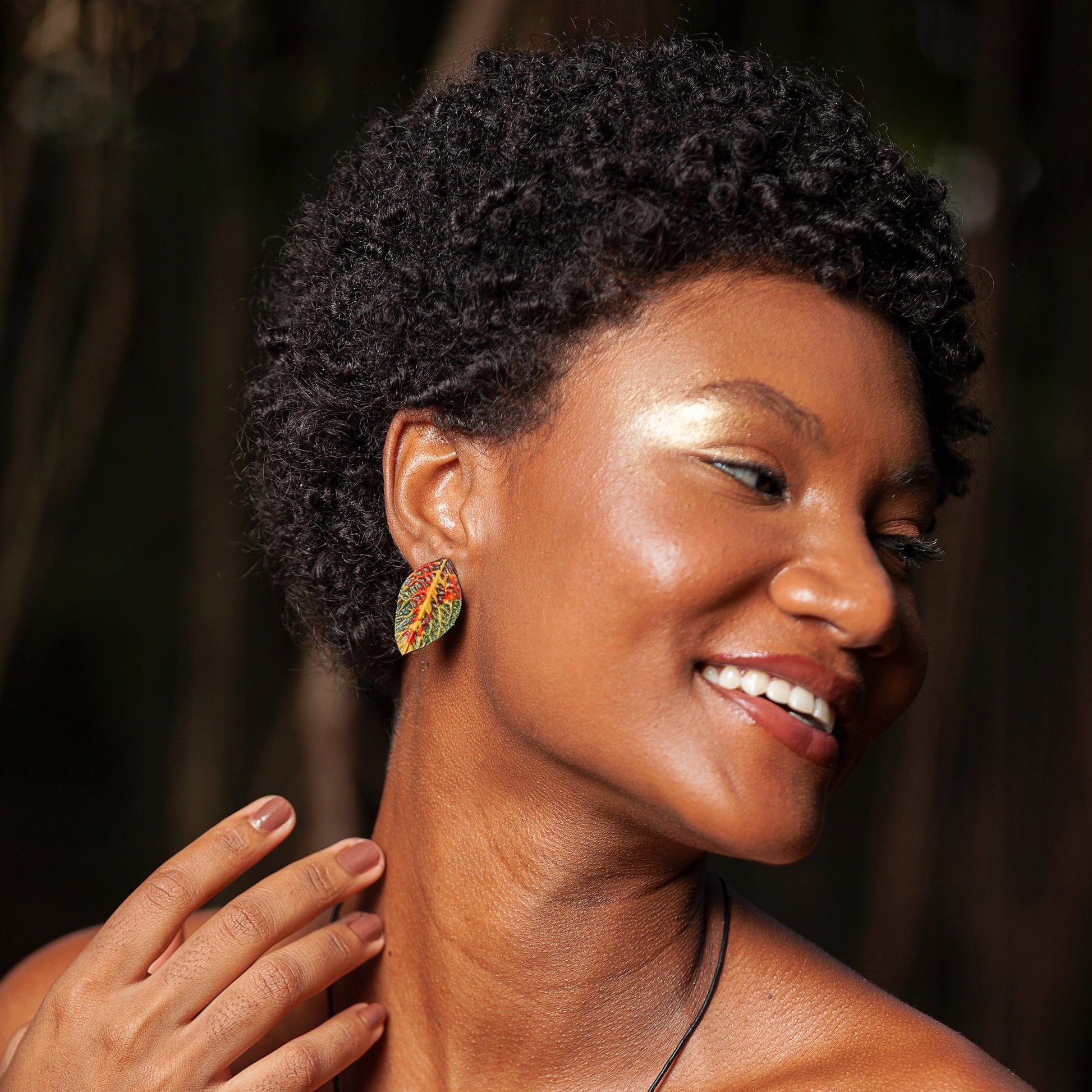 A radiant woman with a joyful smile and curly hair showcases a colorful leaf-shaped earring, complementing her luminous golden makeup and the natural backdrop.