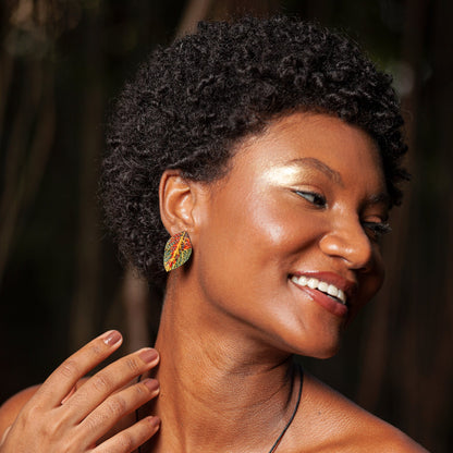 A radiant woman with a joyful smile and curly hair showcases a colorful leaf-shaped earring, complementing her luminous golden makeup and the natural backdrop.