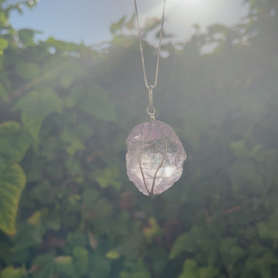 Raw amethyst pendant on a silver necklace. Video shows the pendant swaying with a sunset and green leaf wall background.