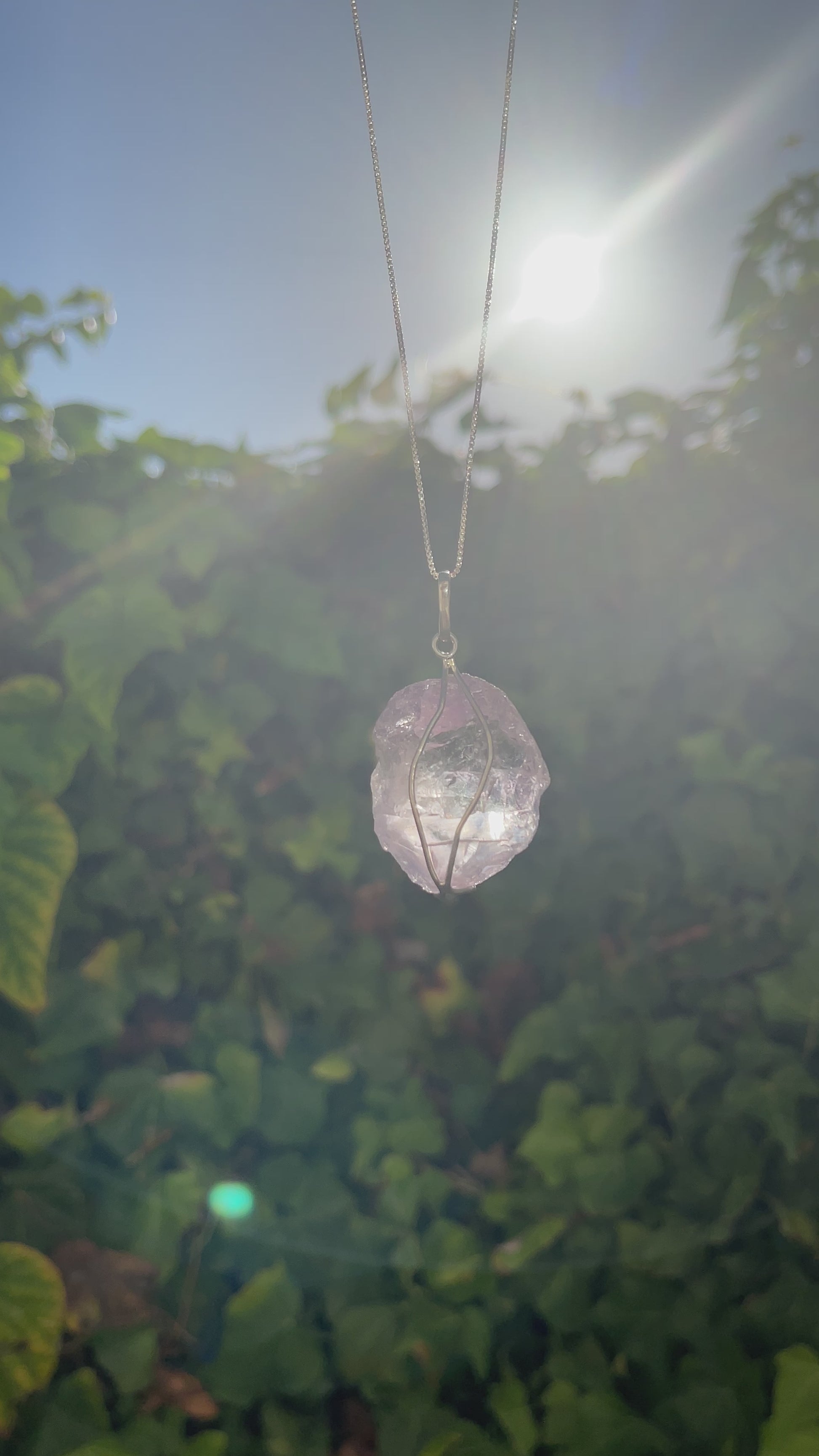 Raw amethyst pendant on a silver necklace. Video shows the pendant swaying with a sunset and green leaf wall background.