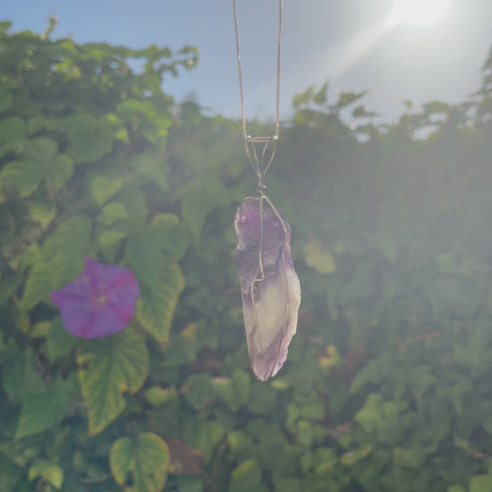 An uncut amethyst gemstone pendant, its violet tones fading into white, suspended by a whimsical heart-crafted silver wire.