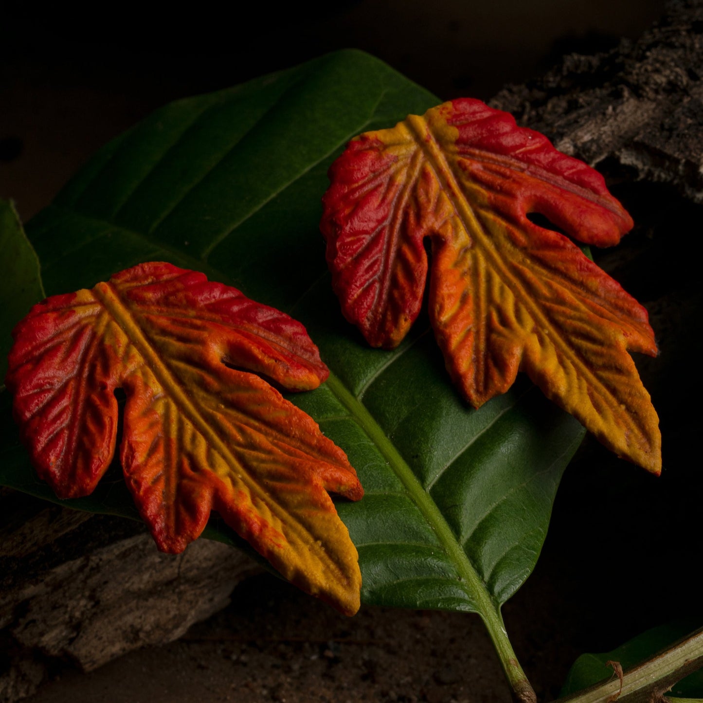 Vibrant orange and yellow leaf-shaped earrings rest on a lush green leaf, both showcasing detailed vein patterns.