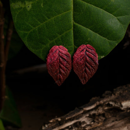 Red leaf-shaped earrings, mimicking the texture of foliage, hang against a green leaf backdrop, blending seamlessly with the natural environment.